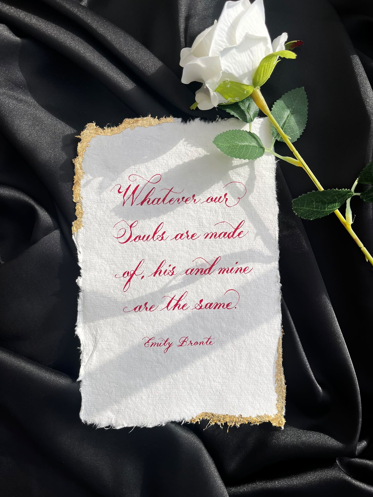 “Whatever our souls are made of, his and mine are the same” by Emily Brontë, hand-lettered in red Copperplate calligraphy on handmade paper with gold-leaf edges, beside a white rose on black satin.