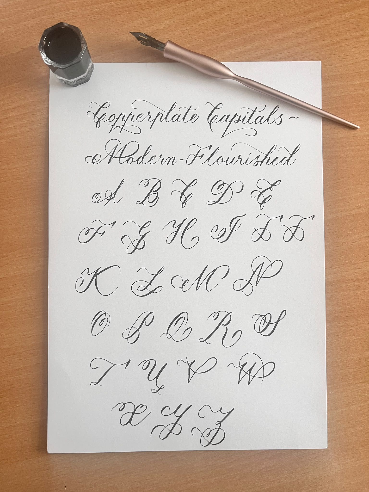 A hand-written Copperplate capitals exemplar headed “Copperplate Capitals ~ Modern-Flourished”, the full flourished alphabet A to Z in black ink on white paper, with a rose-gold oblique holder and a glass pot of ink alongside.