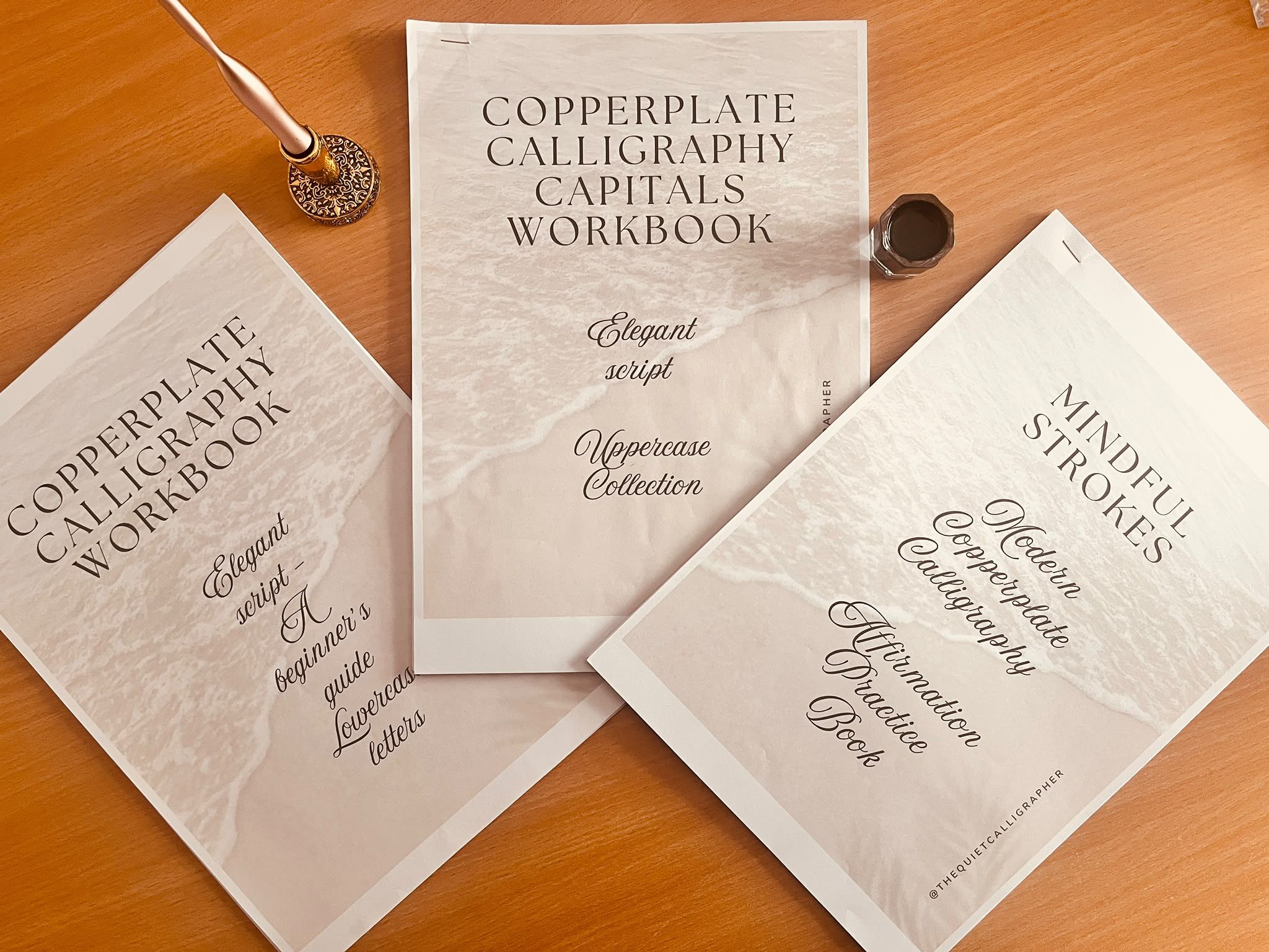 Three printed Copperplate workbooks fanned out on a wooden desk — the Copperplate Calligraphy Workbook, the Copperplate Calligraphy Capitals Workbook and Mindful Strokes — beside an ornate gold pen holder and a pot of ink.