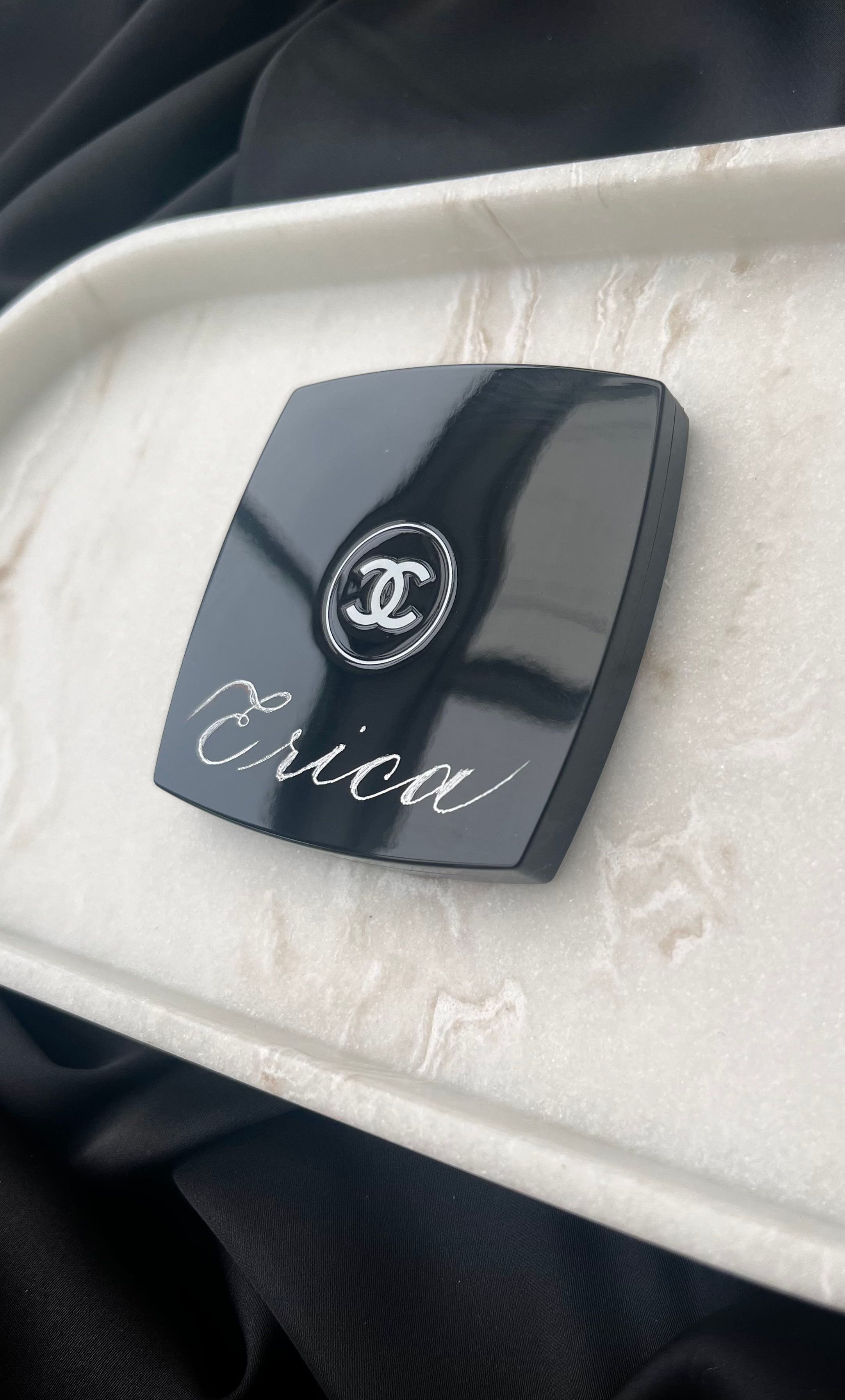 “Erica” hand-engraved in silver Copperplate script across the lid of a black Chanel compact, resting on a white marble tray.
