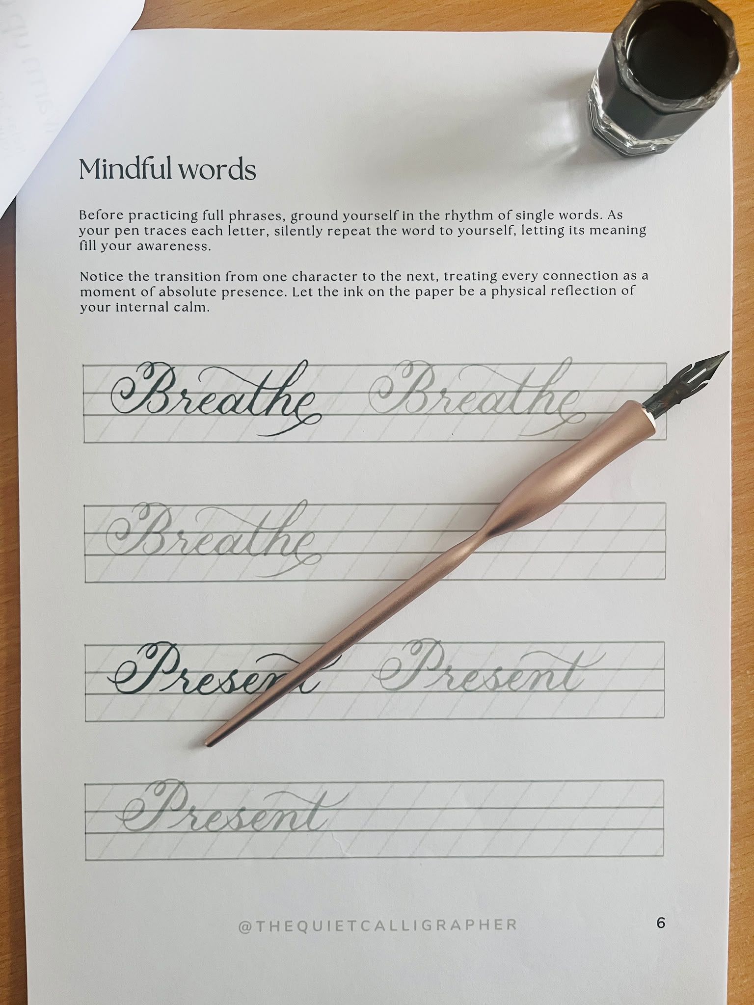 A workbook practice page headed “Mindful words”, with ruled Copperplate guidelines and the words “Breathe” and “Present” written in ink above lighter tracing guides, a rose-gold oblique holder resting across the page.