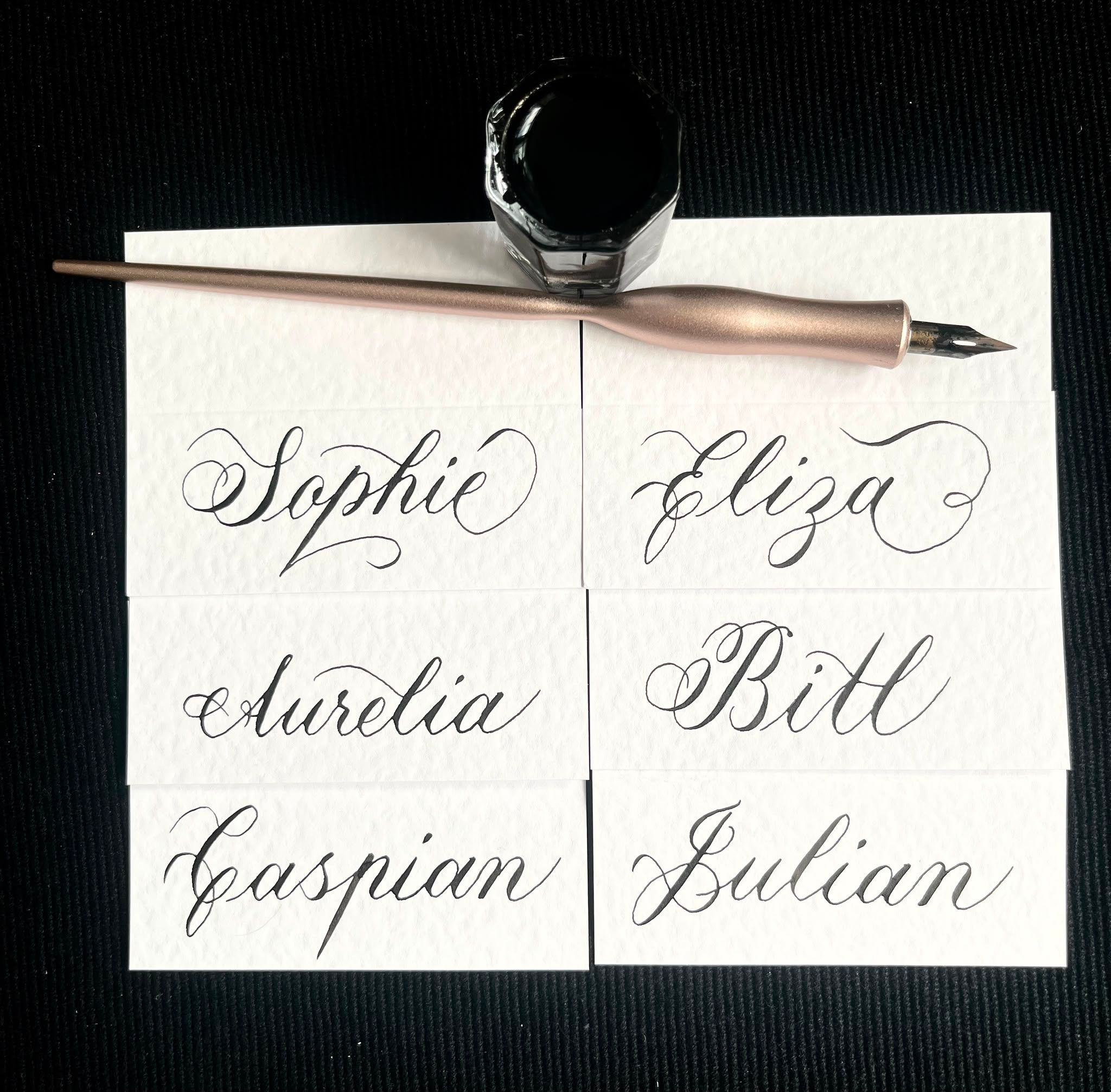 Six place cards hand-lettered in black Copperplate calligraphy (Sophie, Eliza, Aurelia, Bill, Caspian and Julian) beneath a rose-gold oblique pen and a bottle of ink.