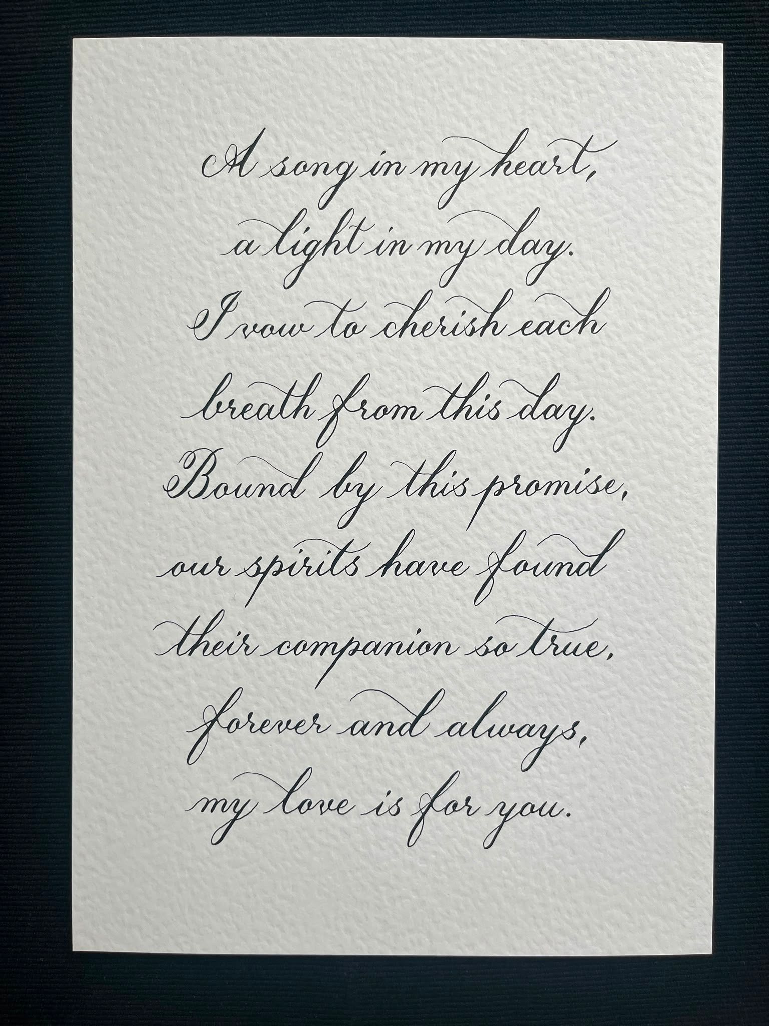 Wedding vows hand-lettered in black Copperplate calligraphy on textured white paper: “A song in my heart, a light in my day. I vow to cherish each breath from this day. Bound by this promise, our spirits have found their companion so true, forever and always, my love is for you.”